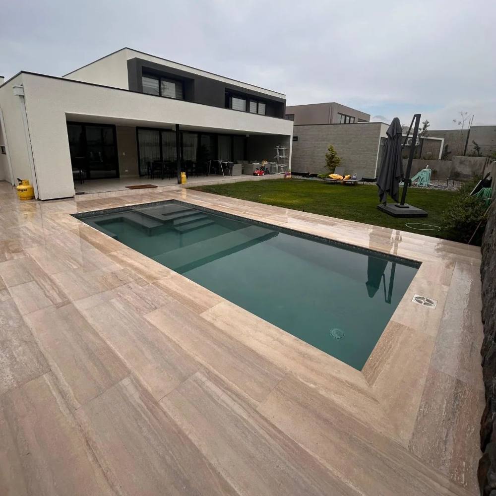 Piscina de hormigón con borde de travertino junto a una casa de dos pisos